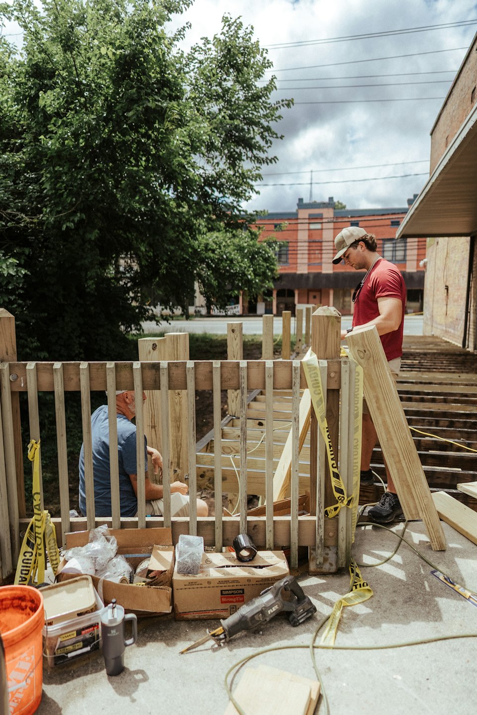 Two carpenters build a wooden deck outdoors near a tree.