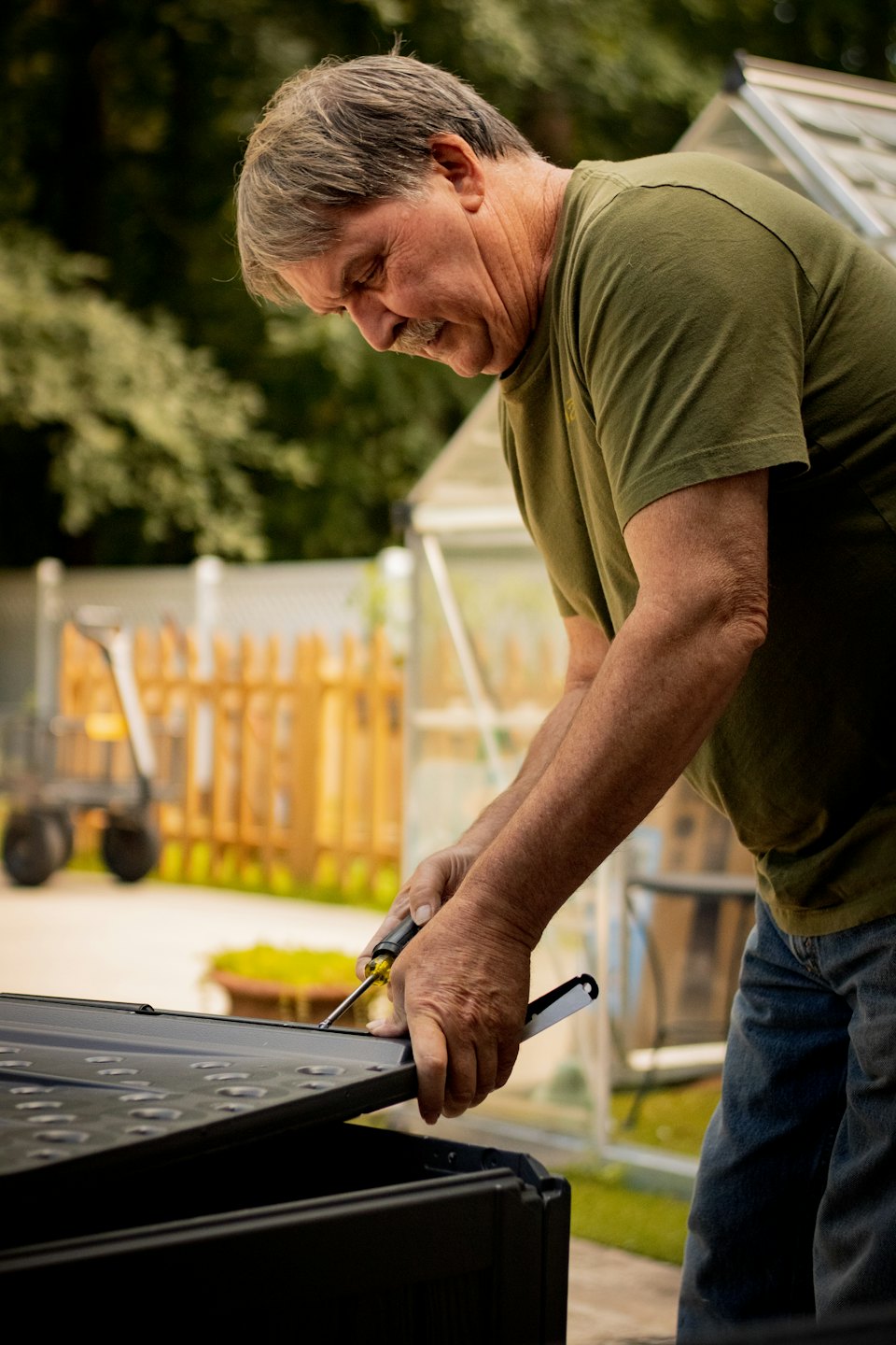 Man in a green shirt uses a screwdriver in a backyard.